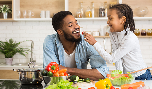 Adorable Black Girl Feeding Her Father With Fresh Tomato In Kitchen, panorama, empty space