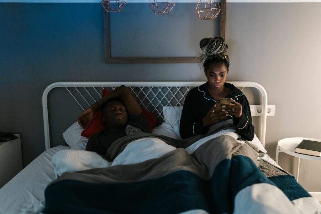 A woman pressing phone on bed beside his sleeping husband 