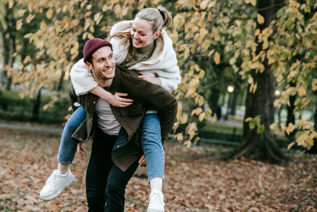 Cheerful man giving piggyback ride to smiling girlfriend in park in autumn